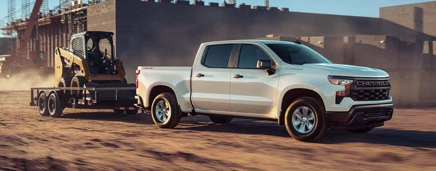 A white 2023 Chevy Silverado 1500 WT is shown towing heavy machinery through a construction site.